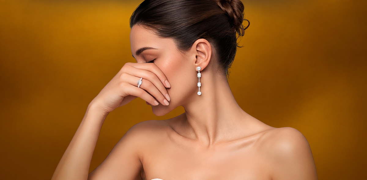 A woman with her hair in a bun wears a delicate silver ring on one finger and a matching silver drop earring with clear stones, set against a warm golden background.