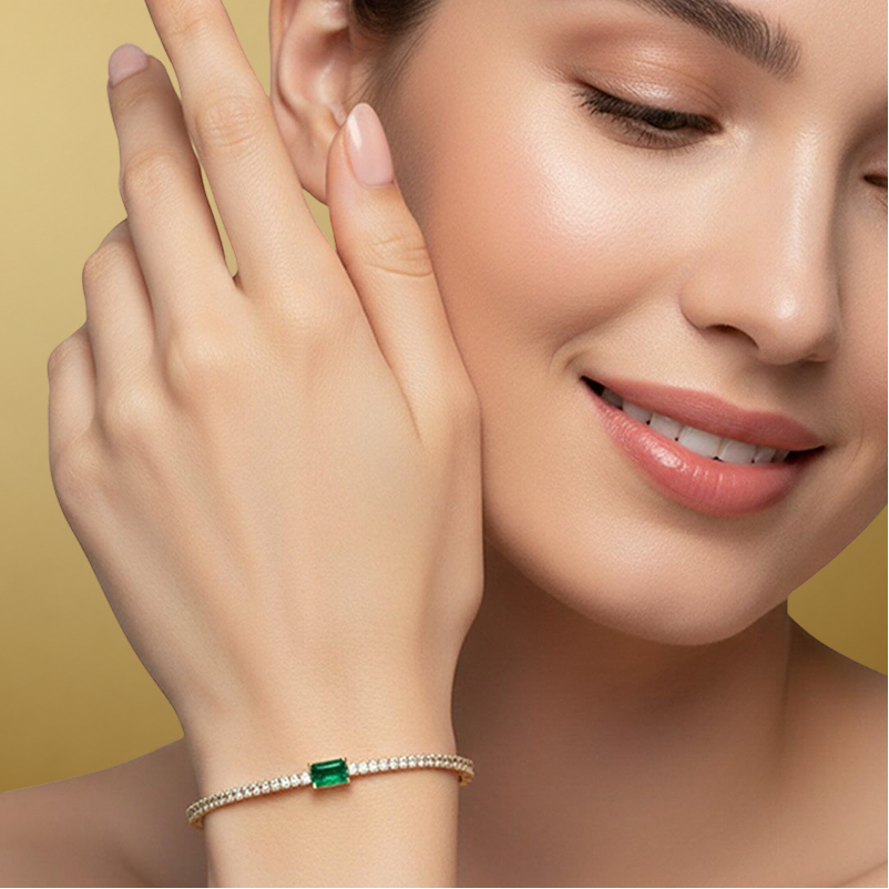 A close-up lifestyle shot of a woman wearing a delicate silver-toned bracelet with a rectangular green emerald-cut center stone and small clear accent stones, set against a soft warm gold background.1
