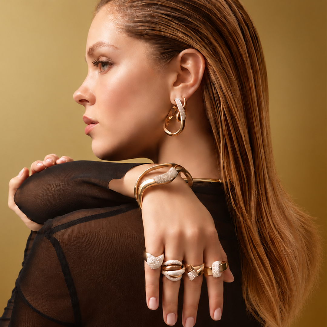 A woman in a sheer black top models gold and silver jewelry, including a large hoop earring, stacked bracelets, and multiple statement rings, against a warm gold background.
