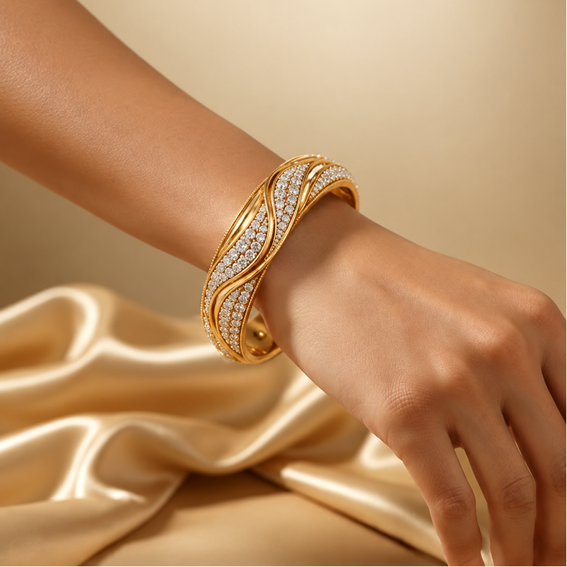 A close-up of a person's hand and wrist wearing a wide gold bangle bracelet with pavé-set clear stones, posed against a warm beige background with draped satin fabric.2